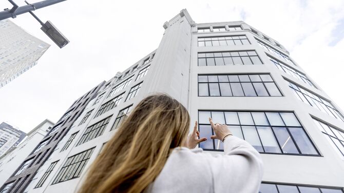 Kobieta unosząca ręce w kierunku białego wieżowca z dużą ilością okien.