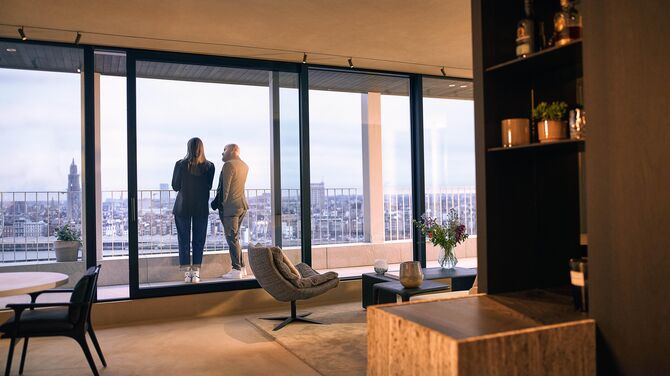 Alt: Widok wnętrza nowoczesnego apartamentu z szerokimi drzwiami przesuwnymi i dwiema osobami na balkonie.