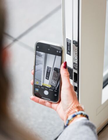 Dłoń trzymająca smartfon blisko kodu QR na białym profilu okna.